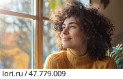 A young woman in a yellow sweater looks out of a window, with sunlight touching her face and curly hair. Стоковое AI-изображение, AI-художник Данила Большаков / Фотобанк Лори