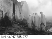 An atmospheric black and white photograph depicting a group of people, wrapped in long white cloths, walking along a ruined wall in the fog. The scene is filled with a sense of mystery, ritual and symbolism. The contrast between the light figures and the dark, blurry surroundings creates a dramatic mood. The photo evokes themes of memory, faith, loss, silence and transition, making it suitable for conceptual and documentary visual content. Стоковое AI-изображение, AI-художник Анна Кривицкая / Фотобанк Лори