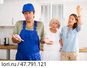 Inattentive plumber in uniform spreading his arms to the sides while being scolded by two women in the kitchen. Стоковое фото, фотограф Яков Филимонов / Фотобанк Лори