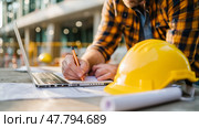 Construction worker reviews plans and takes notes while using a laptop at a job site in a busy urban environment during the day. Стоковое AI-изображение, AI-художник Сергей Толмачёв / Фотобанк Лори