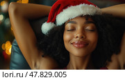 A young woman with a Santa hat smiles contentedly while relaxing in a cozy chair, enveloped by warm holiday lights and decorations. Стоковое AI-изображение, AI-художник Данила Большаков / Фотобанк Лори