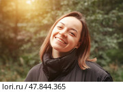 A young woman stands joyfully in a lush forest, her warm smile radiating happiness against the backdrop of vibrant greenery. Sunlight filters through the trees, creating a magical atmosphere. Стоковое фото, фотограф Aleksandr Golubev / Фотобанк Лори