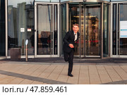 A cheerful man in a suit rushes out of a sleek office building, showing excitement as he exits. The warm glow of sunset highlights his energetic stride and joyful expression in an outdoor setting. Стоковое фото, фотограф Aleksandr Golubev / Фотобанк Лори