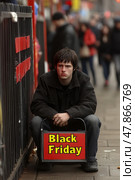 A young man sits on the sidewalk holding a Black Friday sign while passersby shop on a busy city street during the holiday season. Стоковое AI-изображение, AI-художник Данила Большаков / Фотобанк Лори