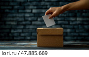 A hand is inserting a ballot into a cardboard box, symbolizing participation in an election, set against a dark brick wall backdrop. Стоковое AI-изображение, AI-художник Данила Большаков / Фотобанк Лори