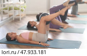 Motivated sporty young girl doing toe taps exercise with Pilates ball between knees while lying on mat, focusing on strengthening core and hip muscles, in group fitness class in sunlit studio. Стоковое видео, видеограф Яков Филимонов / Фотобанк Лори