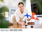 Positive young woman putting flag of Finland on the table with flag of Switzerland in conference room. Стоковое фото, фотограф Яков Филимонов / Фотобанк Лори