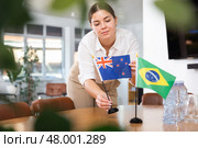 Positive young woman putting flag of Australia on the table with flag of Brazil in conference room. Стоковое фото, фотограф Яков Филимонов / Фотобанк Лори