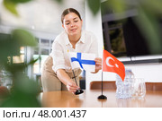 Positive young woman putting flag of Finland on the table with flag of Turkey in conference room. Стоковое фото, фотограф Яков Филимонов / Фотобанк Лори