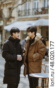 Two young men chat intimately while holding cups on a chilly winter day in a quaint street. Стоковое AI-изображение, AI-художник Данила Большаков / Фотобанк Лори