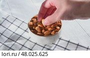 Hand picking an almond from a white ceramic bowl overflowing with almonds, resting on a checkered tablecloth atop a light wooden table, highlighting healthy snacking options. Стоковое видео, видеограф Ольга Ильина / Фотобанк Лори