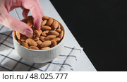 Hand picking an almond from a white ceramic bowl overflowing with almonds, resting on a checkered tablecloth atop a light wooden table, highlighting healthy snacking options, copy space. Стоковое видео, видеограф Ольга Ильина / Фотобанк Лори