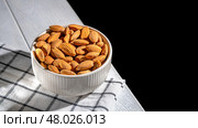 White bowl brimming with almonds resting on a checkered cloth atop a wooden table, creating a cozy, inviting atmosphere against a dark background. Perfect for highlighting healthy snacking. Стоковое фото, фотограф Ольга Ильина / Фотобанк Лори