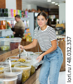 Young girl chooses marinated olives in supermarket, scooping them from bucket with ladle into plastic bag. Стоковое фото, фотограф Яков Филимонов / Фотобанк Лори