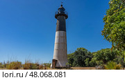 An old lighthouse on a tropical island. A tall black and white cylindrical tower against a clear blue sky. The weather vane is at the top. Green vegetation. Madagascar. Faro Nosy Iranja (2023 год). Стоковое фото, фотограф Вера Ивановна Кубарева / Фотобанк Лори