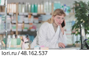 Young woman pharmacist answers a phone call near the computer, she checks the availability of medicines and pills for a client. Pharmacy employee advises the client by phone. Стоковое видео, видеограф Яков Филимонов / Фотобанк Лори