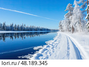 Serene view of the river with a path on bank along trees with blue sky and contrail on sunny winter day. Стоковое AI-изображение, AI-художник Данила Большаков / Фотобанк Лори