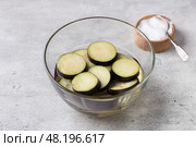 Soaking eggplant slices in salt water in a glass bowl on a gray textured background. Processing eggplant before cooking, removing bitterness. Стоковое фото, фотограф Ольга Лепёшкина / Фотобанк Лори