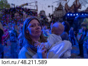 A woman embraces a baby as confetti falls during an evening festival filled with joyful crowds and vibrant lights. Стоковое фото, фотограф Данила Большаков / Фотобанк Лори