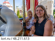 A mother holds her baby while smiling on a carousel, surrounded by vibrant colors and other playful attractions. Стоковое фото, фотограф Данила Большаков / Фотобанк Лори