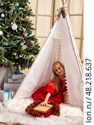 A six-year-old girl model in a vibrant red dress is nestled inside a cozy teepee. Surrounded by a beautifully decorated Christmas tree, she holds a whimsical book while festive gifts await nearby. Стоковое фото, фотограф Aleksandr Golubev / Фотобанк Лори