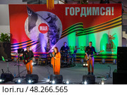 Концерт музыкальной группы «План Б» во время праздничного мероприятия «День Победы на века!», посвящённого 80-летию Победы в Великой Отечественной войне. Спорткомплекс «Президентский», город Магадан, Магаданская область, Россия. На сцене музыканты и певцы исполняют песни военных лет. На стене баннер с надписями «9 мая», «Гордимся!». Редакционное фото, фотограф Андрей Степанов / Фотобанк Лори