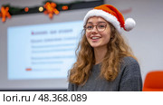 A cheerful young woman in a Santa hat radiates joy while participating in a holiday presentation at school. Стоковое AI-изображение, AI-художник Данила Большаков / Фотобанк Лори