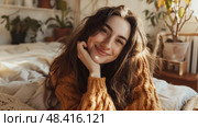 A young woman with long brown hair, wearing a yellow sweater, relaxes on a bed with a white comforter. She smiles as she looks at the camera. Стоковое AI-изображение, AI-художник Данила Большаков / Фотобанк Лори