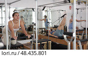 Positive concentrated young girl practicing pilates stretching exercises on reformer at gym. Стоковое видео, видеограф Яков Филимонов / Фотобанк Лори