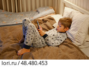 Cute kid boy in pajamas lying in bed watching smartphone, smiling. Children's screen time. Child plays games using phone at the evening or night. Social media addiction. Childhood, modern technology. Стоковое фото, фотограф Марина Демидюк / Фотобанк Лори