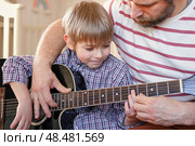 Handsome dad teaches his cute son to play the guitar sitting on sofa in the living room at home. Happy Father's Day. Father teaching his child to play guitar. Kid Learning music skill. Art education. Стоковое фото, фотограф Марина Демидюк / Фотобанк Лори