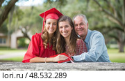 A happy graduate in red celebrates with her supportive family, embracing outdoors on a sunny day. Стоковое AI-изображение, AI-художник Данила Большаков / Фотобанк Лори