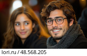 A young man with glasses smiles warmly, dressed in a dark scarf, as a woman gazes at him with a smile in a lively gathering. Стоковое AI-изображение, AI-художник Данила Большаков / Фотобанк Лори