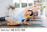 Elderly woman in company of hatha yoga lovers performs half-lotus pose, Ardha Padmasana. Стоковое фото, фотограф Яков Филимонов / Фотобанк Лори