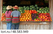 At a lively market in the countryside, two women in traditional clothing choose from an array of colorful fresh fruits on display. Стоковое AI-изображение, AI-художник Данила Большаков / Фотобанк Лори
