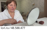 A beautiful senior woman is sitting at home and doing her make-up morning routine. Senior woman putting blush on her face. Стоковое видео, видеограф Ирина Старикова / Фотобанк Лори