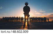 A silhouetted figure of an officer stands in the foreground with a blazing sunset behind, facing a line of uniformed personnel. Стоковое AI-изображение, AI-художник Данила Большаков / Фотобанк Лори