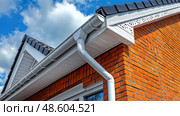 A detailed look at the eaves and gutters of a contemporary brick home with clouds in the sky. Стоковое AI-изображение, AI-художник Данила Большаков / Фотобанк Лори