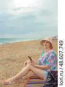 Woman in sun hat and bright blouse sitting on beach, holding phone and mysteriously looking at sea. Стоковое фото, фотограф Коханова Наталья / Фотобанк Лори