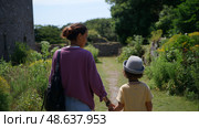 Mother and child walking hand in hand on a sunny day in a green landscape near ancient stone ruins. Стоковое AI-изображение, AI-художник Сергей Толмачёв / Фотобанк Лори