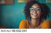 A cheerful young woman, with curly hair and glasses, smiles contentedly while sitting against a colorful backdrop in a cozy indoor setting. Стоковое AI-изображение, AI-художник Данила Большаков / Фотобанк Лори