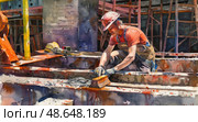 A construction worker is focused on applying cement to a structure at a bustling construction site under bright sunlight. Стоковое AI-изображение, AI-художник Данила Большаков / Фотобанк Лори