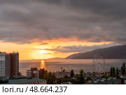 Golden sunlight bathes the coastline as clouds linger over the mountains, with a ferris wheel standing on the shore, evoking a serene atmosphere, Гагра, Абхазия (2023 год). Редакционное фото, фотограф Данила Большаков / Фотобанк Лори