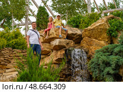 Two children relax on rocks near a waterfall, while an adult stands nearby in a lush, vibrant garden during a sunny summer day, Главрыба, Республика Дагестан, Казбековский район, село Ново-Зубутли. Стоковое фото, фотограф Данила Большаков / Фотобанк Лори