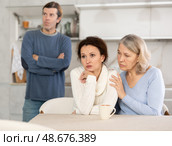 Man quarrels with his wife in kitchen, standing behind her, and elderly mother hugs and calms her daughter. Стоковое фото, фотограф Яков Филимонов / Фотобанк Лори