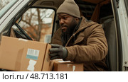 A man in warm clothing carefully unloads packages from a delivery van while seated inside in a chilly residential area. Стоковое AI-изображение, AI-художник Данила Большаков / Фотобанк Лори