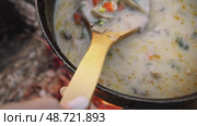 Delicious vegetable soup with mushrooms and cheese is being whisked in camp pot with wooden spoon while cooking over an open flame during hiking expedition. Стоковое видео, видеограф Strunov David / Фотобанк Лори