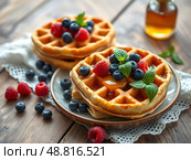 Belgian waffles with honey and fresh blueberries, raspberries and mint leaves on a wooden table with a beautiful napkin. Delicious dessert. Copy space. Стоковое AI-изображение, AI-художник Iva Noel / Фотобанк Лори
