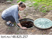 A woman looks into an open septic tank to check the level of sewer discharge. Septic tanks in a country house. Стоковое фото, фотограф Коханова Наталья / Фотобанк Лори