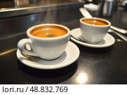 Two Espresso cups with coffee. Freshly brewed espresso shots on a dark wooden table with copy space, background, caffeine. Espresso cups, spoons, saucers. Refreshing hot cup of strong coffee at a cafe. Стоковое AI-изображение, AI-художник Марина Демидюк / Фотобанк Лори